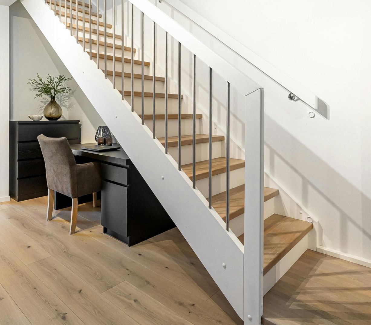 white staircase with light wood treads over a light wooden floor. Black desk and cabinet with light brown chair underneath staircase.