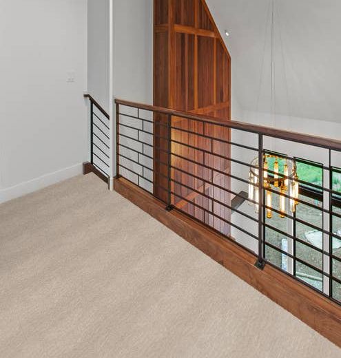 Alistair Stone colored carpet in hallway with white walls and a wooden accent wall with black stair railing.