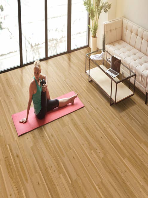 Woman on pink yoga mat stretches beside floor-to-ceiling windows, glass coffee table, and cream sofa