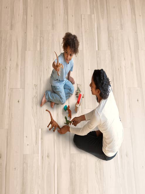 Mother and child play on light beige woodgrain laminate and play with toy dinosaurs