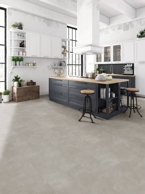 Industrial-inspired white and blue kitchen with large windows, hanging wall cabinets, rustic chest, potted houseplants, and two stools, and light gray stone concrete-look LVT