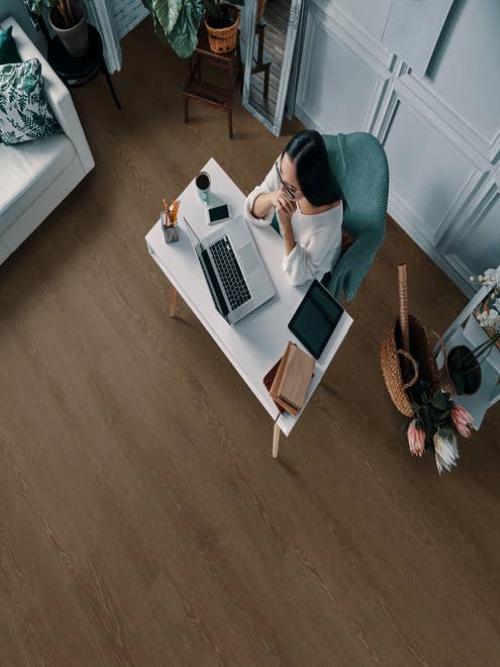 Woman works on laptop on desk in home office, beside sofa, basket of flowers, and shelf, with wainscoting up walls and medium brown woodgrain flooring