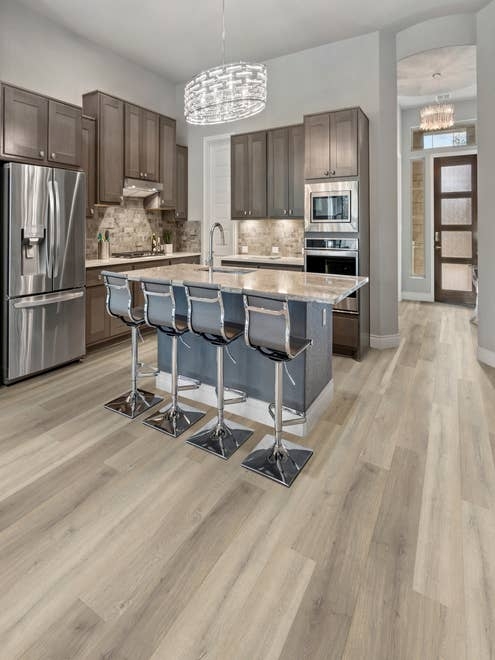 Kitchen with neutral brown cabinets, pale blue accent island with chairs, with wide neutral brownish gray woodgrain flooring, and entry door visible in background