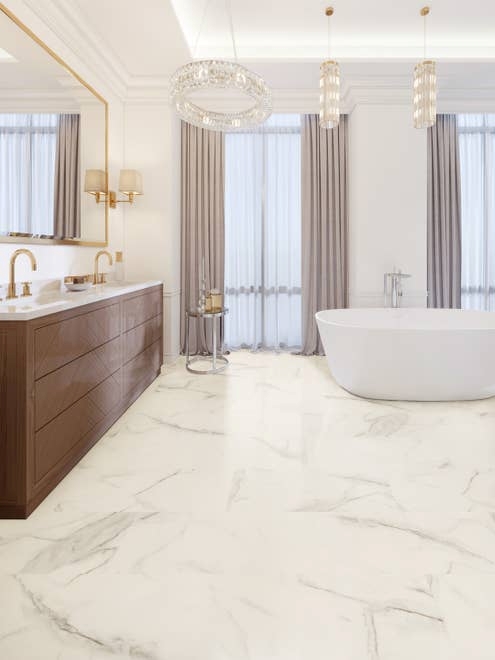 Luxury bathroom with tall windows, multiple chandeliers, tub, wooden vanity, gold mirror and fixtures, and white-and-gold marble stone LVT floor
