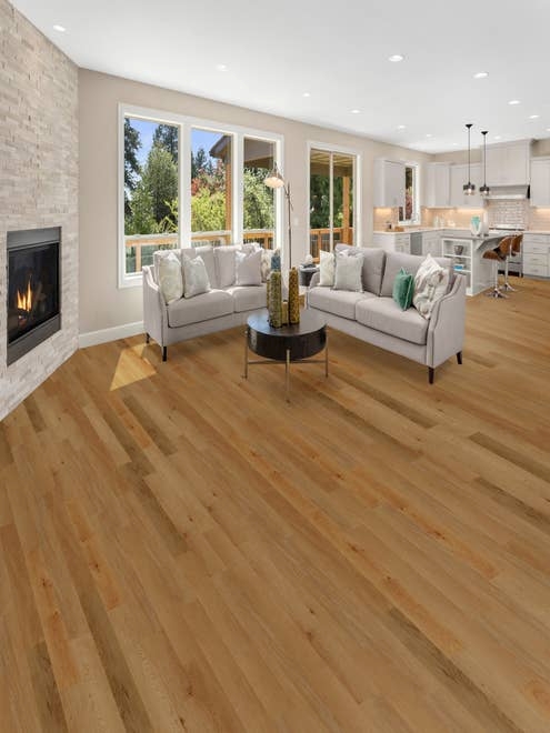 Open-concept kitchen and living room with two sofas and white stone fireplace, with large windows showing pergola, deck, and woods