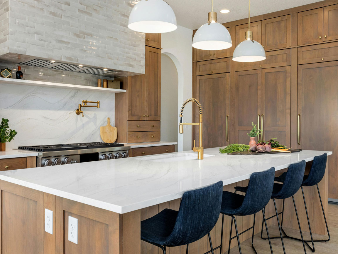 Kitchen cabinetry, dark colored wooden cabinets with gold hardware, silver appliances, blue cloth chairs, white marble counter