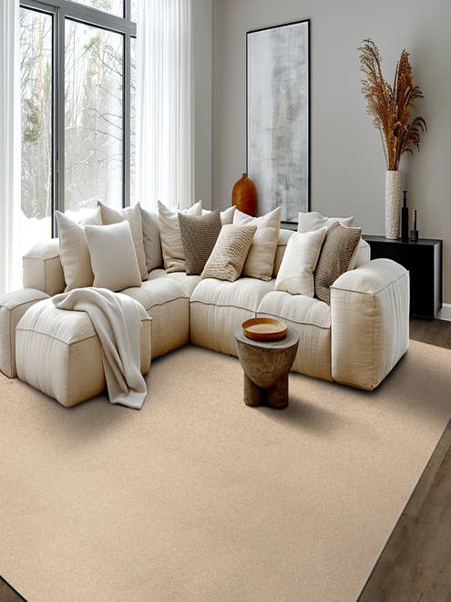 Cozy living room scene with a modern plush cream couch topped with an array of pillows and a throw blanket while snow falls outside