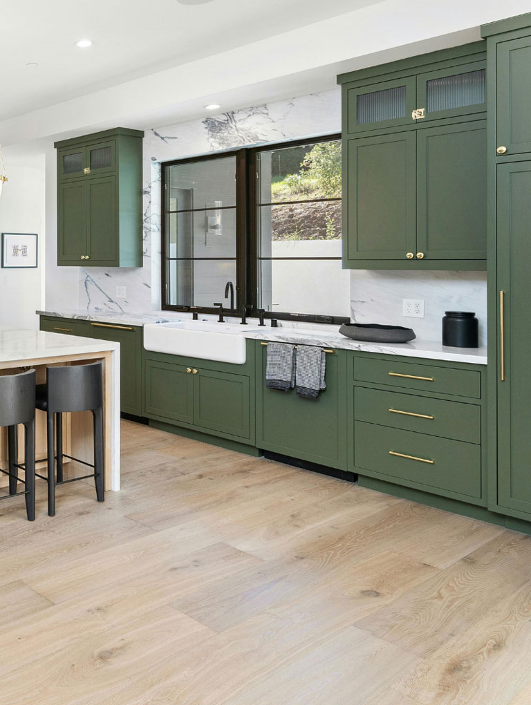 Green kitchen cabinets floor to ceiling with gold hardware, white farmers sink with black faucet, light wood floor, white ceiling and walls