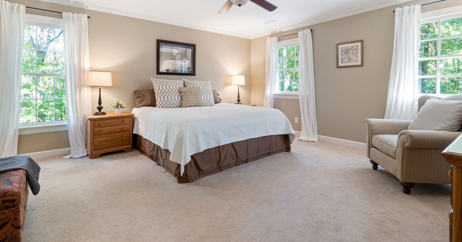 Bedroom with a cream colored carpet, tan walls, tan chair, bed with white comforter, brown bed skirt and wooden bedside table