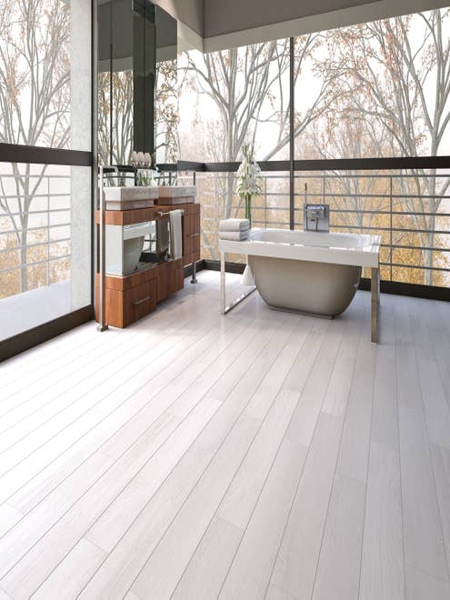 Spa space with bathtub, wooden vanity and mirror, in space with white-gray woodgrain floor and large windows overlooking woods