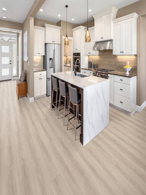 Small modern white kitchen with marble-topped island, seating, with view of entry hall, over light gray woodgrain laminate