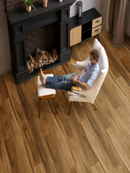 Man in modern, organic armchair sits with feet up on ottoman across from black fireplace, in room with dramatic and rich hickory-look floor