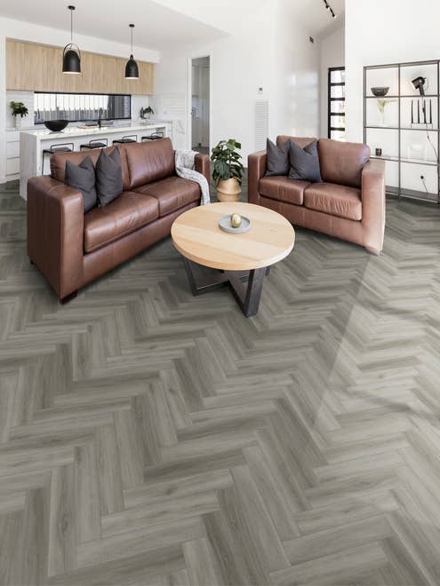Modern open concept living area and kitchen, featuring light/medium gray herringbone floor, leather couches, and white walls