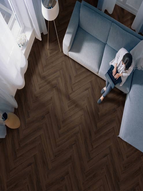 Woman on laptop sits on blue couch in dark and moody living room with dramatic, rich brown herringbone floors