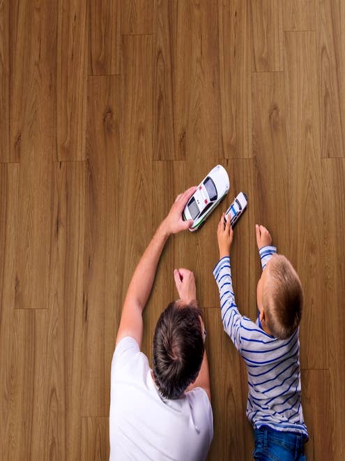 Man and boy lay on rich, reddish brown laminate floor and play with toy cars