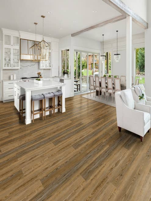 Open-concept kitchen, dining, and seating areas, with white walls, gold accents, and brown naturally-aged luxury vinyl plank floor