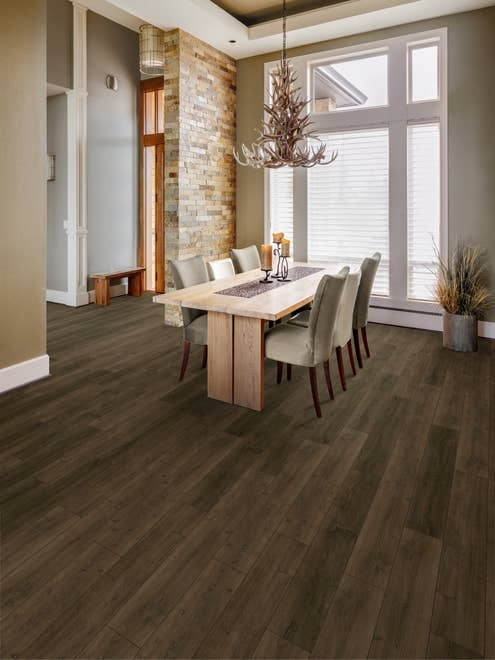 Dining area with table and chairs beneath antler chandelier, in room with exposed brick wall, large window, and entryway in background, with medium to dark brown LVP flooring