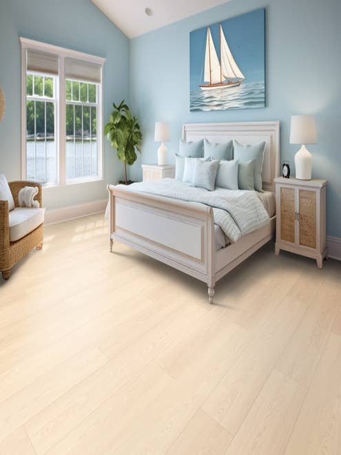 Bright, airy bedroom with bed, nightstand, corner chair, potted plant, and sailboat painting with light blue walls and pale woodgrain floor