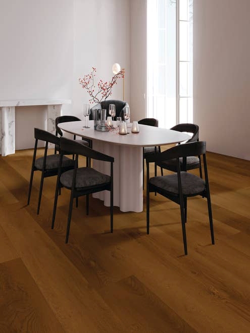 Black chairs and white dining table with glassware and plant sprig in room with large window, medium brown floors, and marble table in background