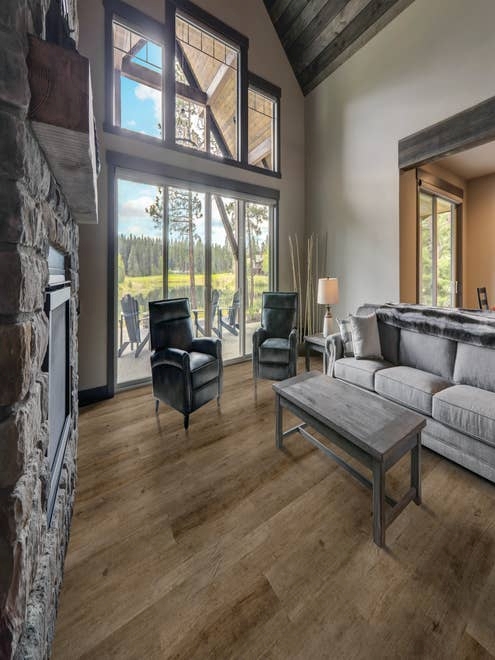Cabin with large fieldstone fireplace, two armchairs, plush gray sofa, and coffee table, with large windows overlooking patio with Adirondack chairs