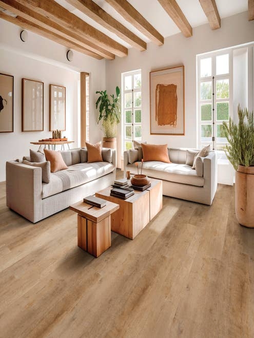 Bright living room with large windows, artwork, two plush sofas facing a coffee table, and houseplants, with exposed wood beams and a tan woodgrain floor