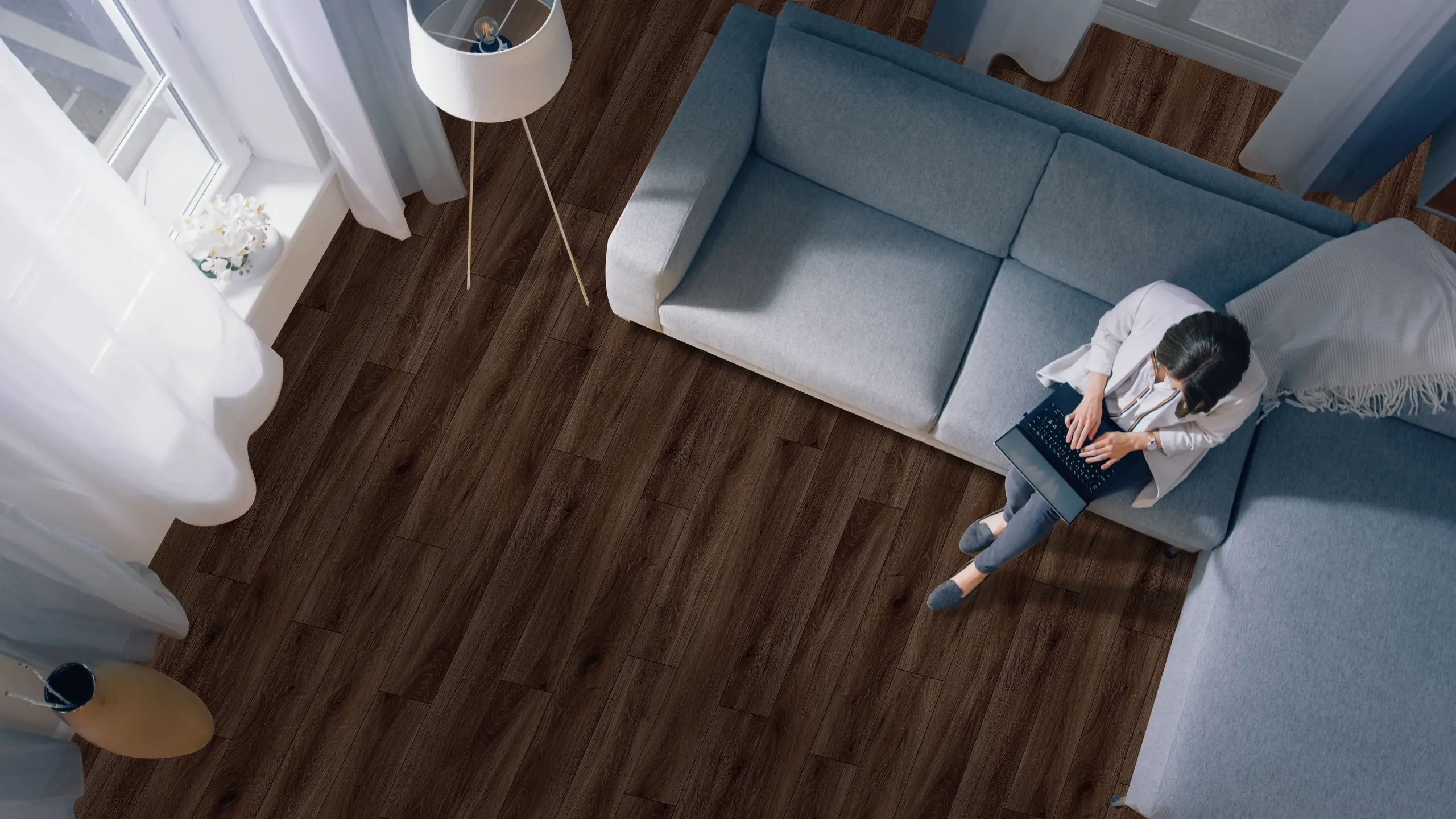 Woman sits on couch and works on laptop beside floor lamp, in calming room with blue walls and rich walnut-color LVP floor