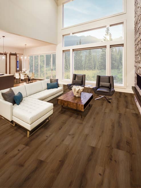Contemporary open concept living room with medium brown wood-look floors with white sectional sofa, two armchairs, coffee table, large stone fireplace, and several large windows showing wilderness beyond