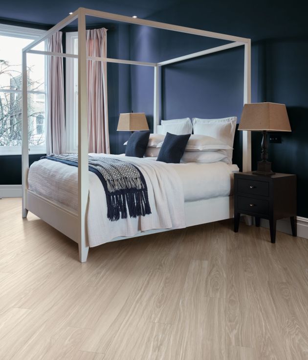 Calm bedroom with dark blue walls, gray woodgrain floors, cozy bed, night stands, large plant, and window