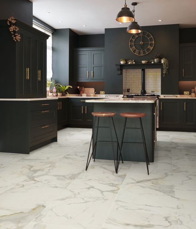 Kitchen with black cabinetry, white counters, and white marble-look tiles, with island with two barstools under pendant lights and wall clock