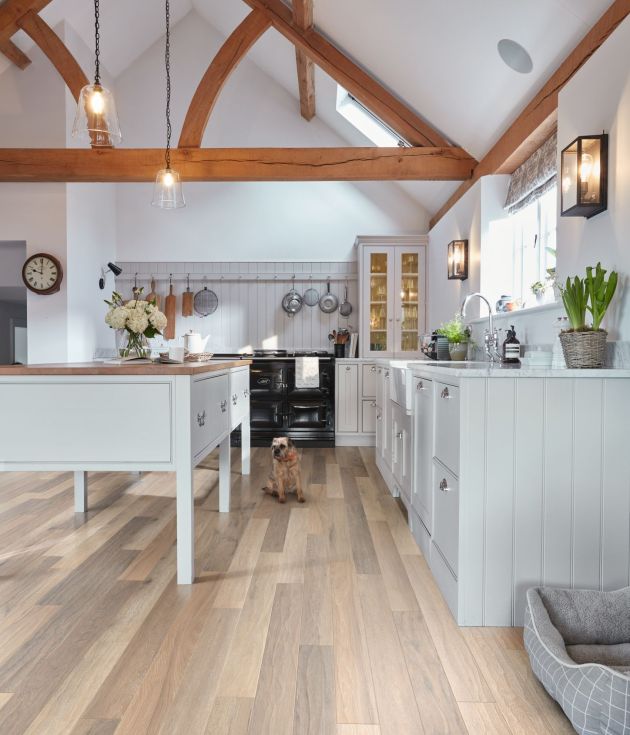 Kitchen with brown woodgrain floor, and white cabinetry, island, with exposed arched wooden beams, and various cooking equipment hanging on wall, with dog sitting before stove