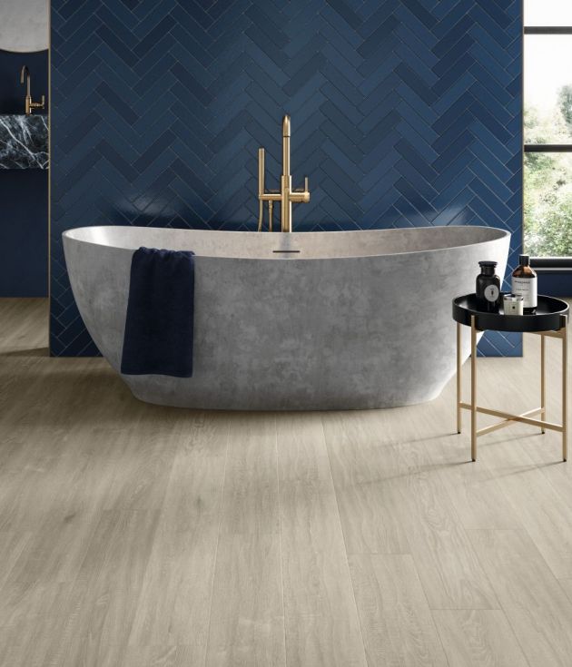 Moody bathroom with taupe woodgrain floor, with large gray soaker tub and table before dark blue herringbone tile wall, with window, potted plant, and sink in background