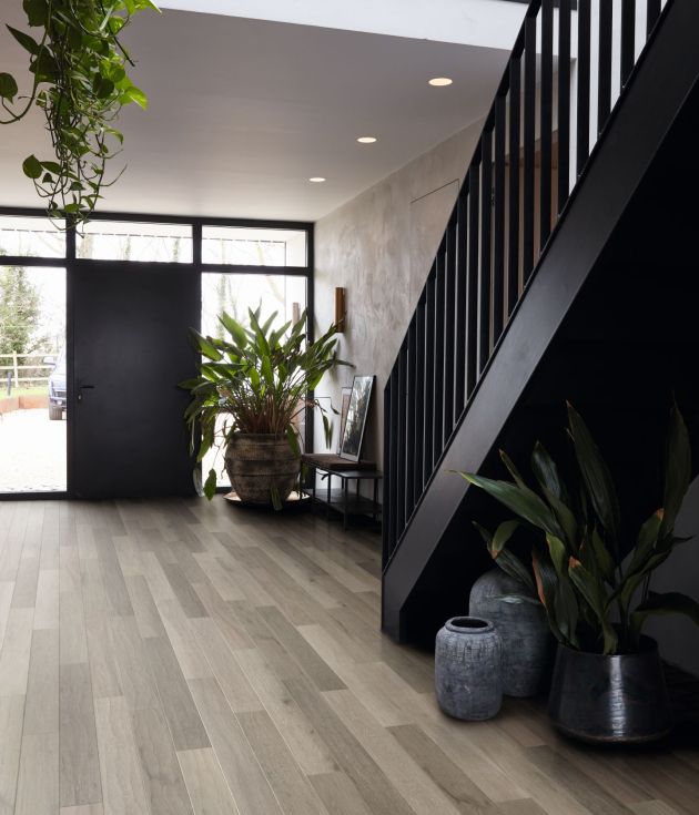 Dramatic entry hallway featuring black staircase, black door surrounded by windows, limestone washed walls, gray woodgrain floor, and several large plants and ceramic vessels, with recessed lighting and glimpse of upper floor