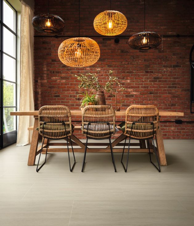 Modern dining table and wicker chairs under multiple woven light fixtures, with exposed brick wall, large black trimmed windows flanked by curtains, iron spiral staircase, and Travertine stone LVT floor