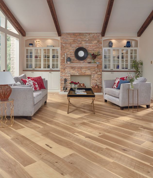 Bright living space with exposed wooden beams, brick fireplace, white built-in cabinets, large window, and contemporary sofa facing coffee table and armchair, with gold metal end tables, over distressed wide-plank hickory woodgrain floor