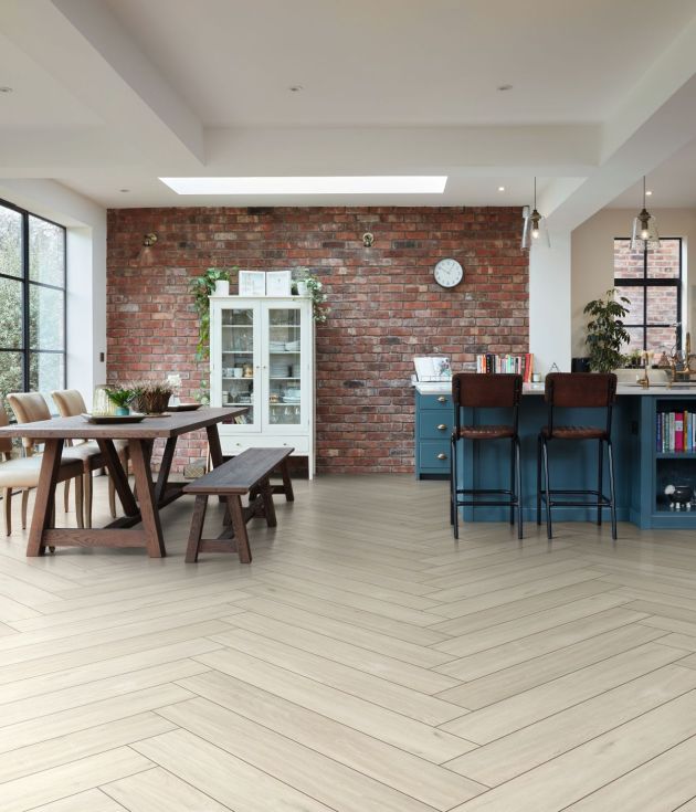 Open-concept kitchen with blue cabinetry, island, and open shelving, and pendant lighting, leading to dining area with table, bench, and chairs before exposed brick wall with China cabinet and large windows, and cream herringbone woodgrain floor