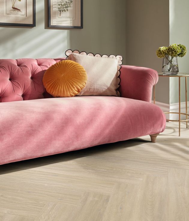 Pink velvet tufted couch and cushions beside gold metal end table, under picture frames, with herringbone woodgrain floor