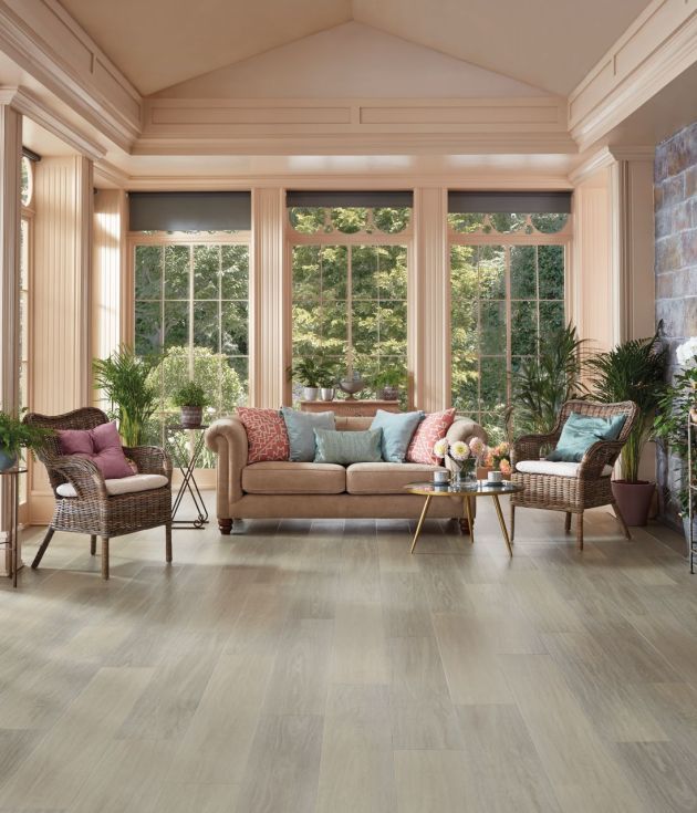 Sunroom with grand windows between columns, exposed brick wall, sofa and wicker chairs, and shelf filled with potted plants, with various other potted plants, with clean gray woodgrain floor
