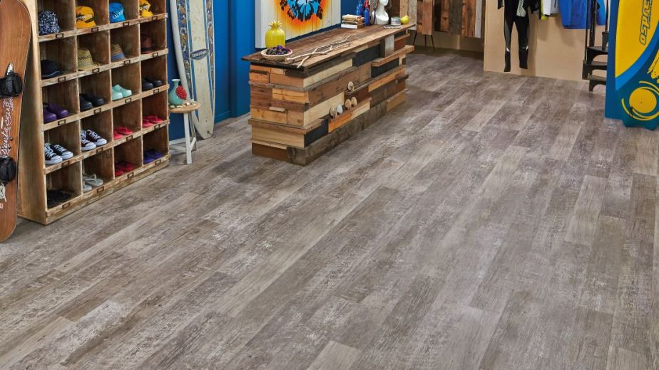 Retail surf shop with shoe cubby shelving, distressed wooden counter, tropical artwork, changing area, and surfboards and surf gear along walls, with gray weathered woodgrain floor