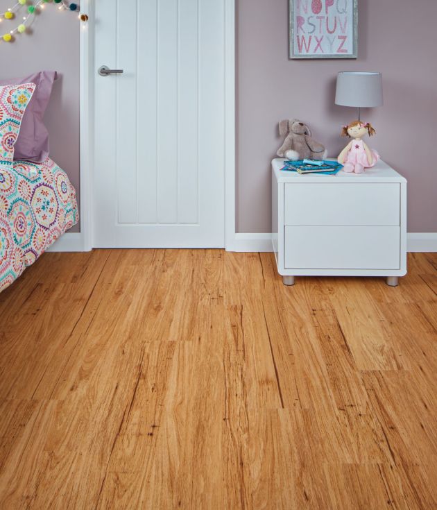Girl's bedroom with lilac walls, white door, golden woodgrain floor, vibrant floral bedspread, and white nightstand with stuffed animals and lamp under alphabet artwork