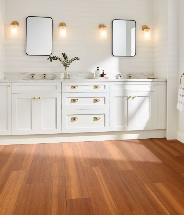 White bathroom dual sink vanity with gold fixtures, sconces, and window with sunlight shining on orange and brown woodgrain floor