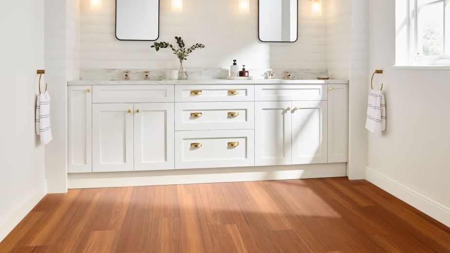 White bathroom dual sink vanity with gold fixtures, sconces, and window with sunlight shining on orange and brown woodgrain floor