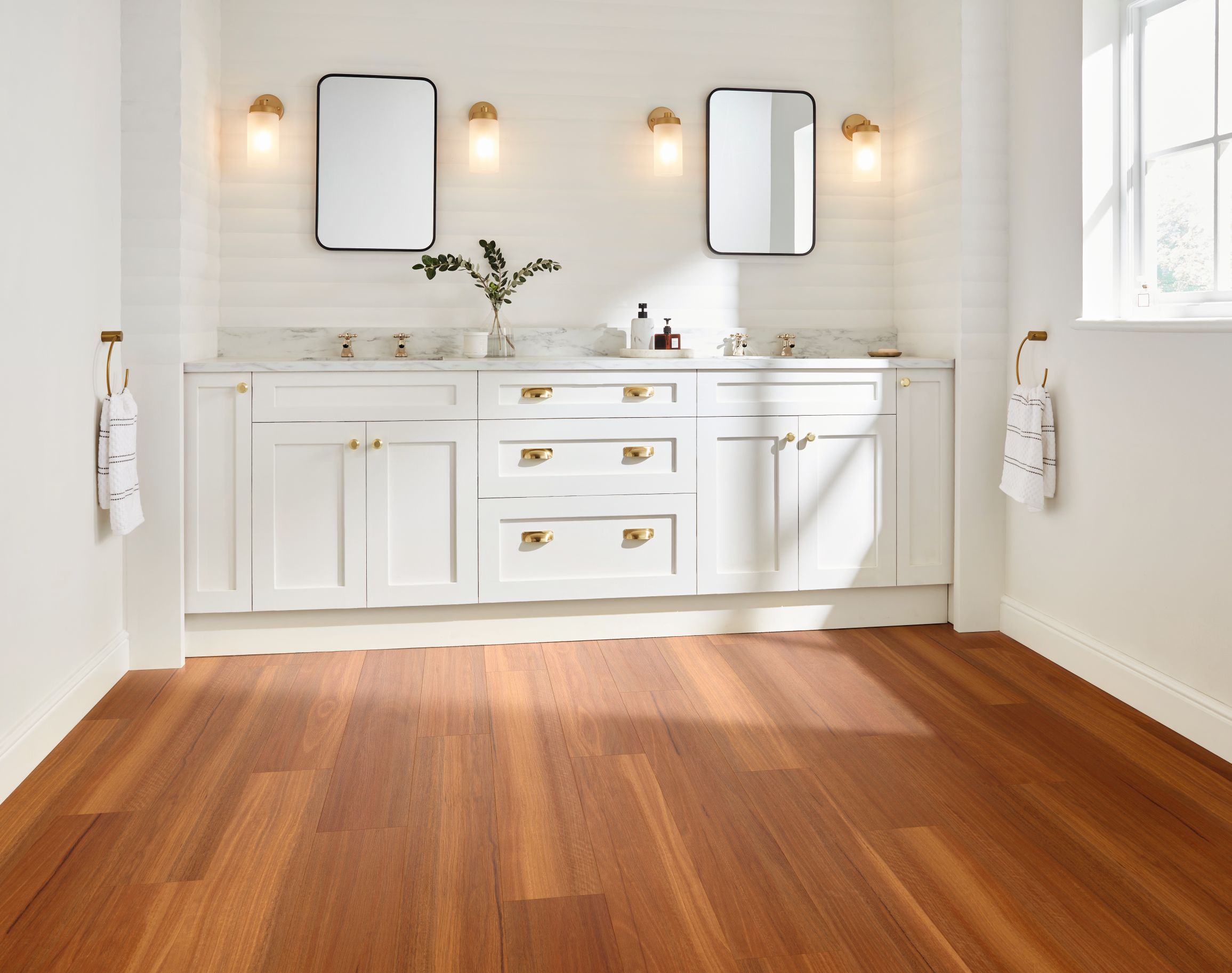 White bathroom dual sink vanity with gold fixtures, sconces, and window with sunlight shining on orange and brown woodgrain floor