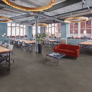Open-concept commercial office space with gray tile floors, oversized light fixtures, meeting desks, red velvet couch, and coffee table, with view of brick building outside