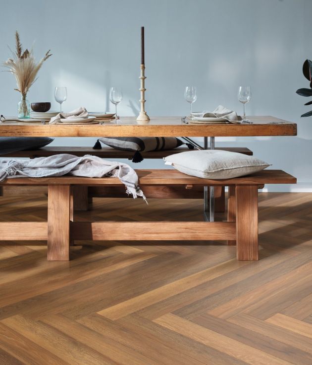 Wooden dining table and bench with wine glasses, candlestick, and pillows over herringbone pattern golden brown LVP floor, with potted plant in corner
