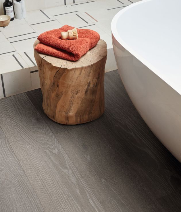 Bathtub and stump side table topped with towel and shave brush, with white and gray geometric shower floor in background, with medium gray woodgrain floor