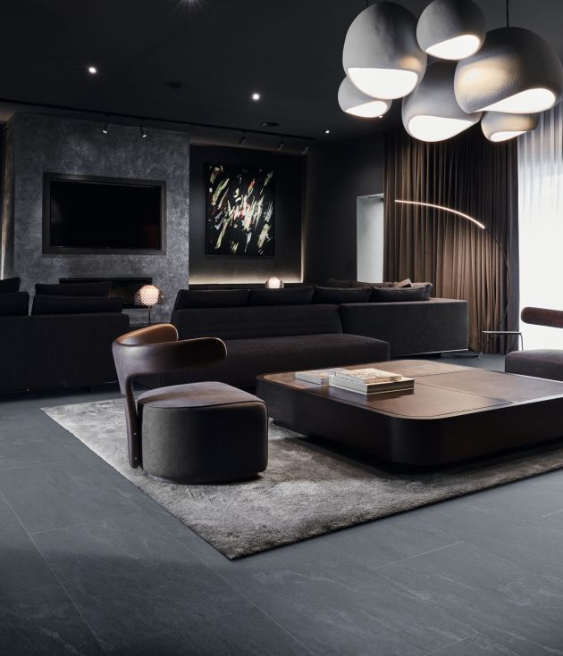 Dark, bold modern living area with large chandelier, TV, and artwork, with couch and chairs around coffee table on area rug and dark tile floor