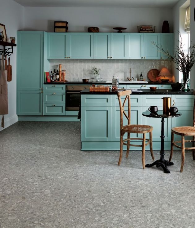 Icy blue kitchen cabinetry and island facing small eating area, with floating shelf and various paraphernalia around, with gray terrazzo-like stone tile floor