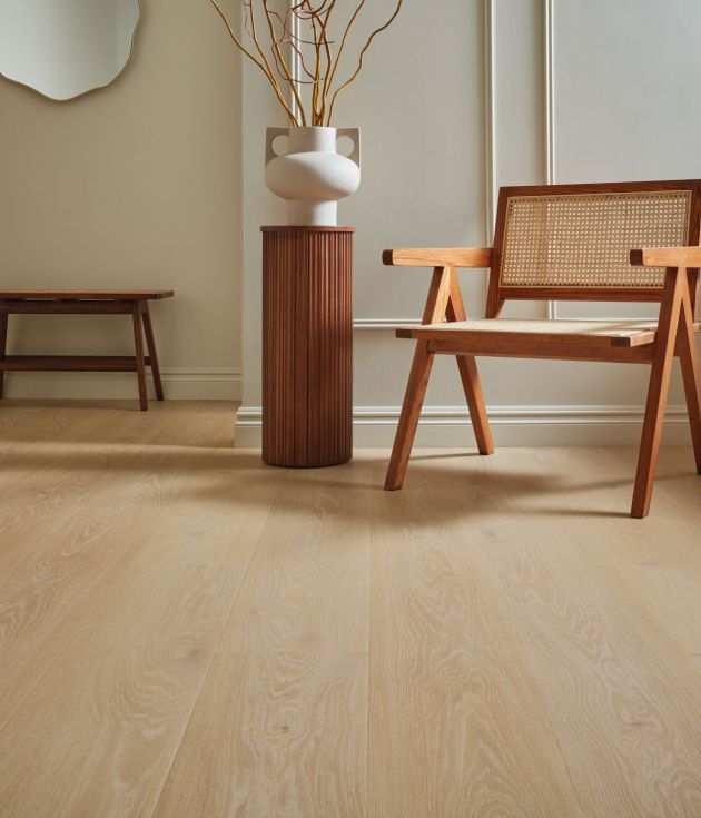 Wooden chair and small wooden cylindrical side table topped with pottery and dried branches, with small wooden bench and mirror in background, in living area with wainscoting and taupe wood-pattern floors