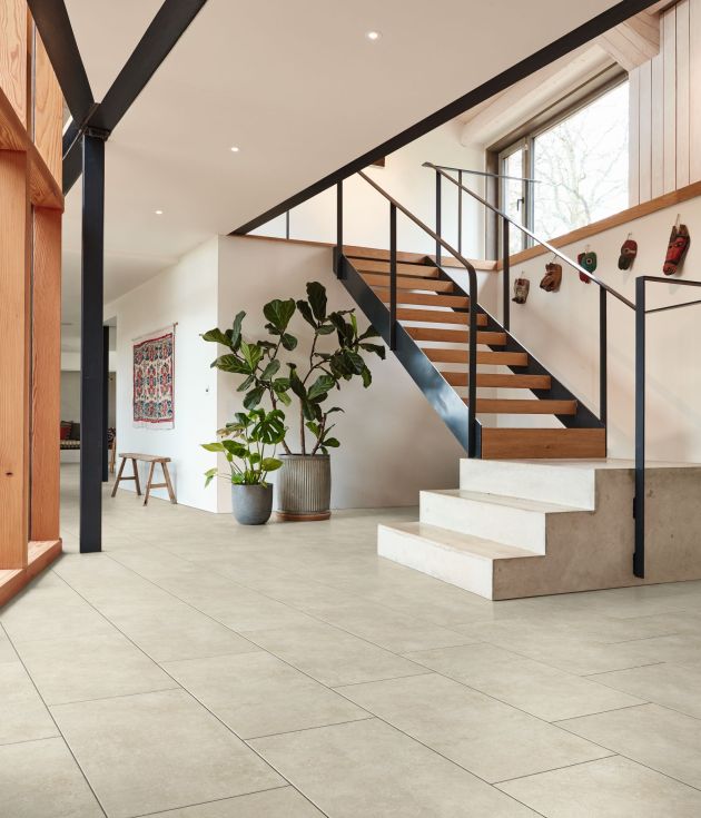 Modern living space and hall with stone steps leading to open wood and metal staircase, various hanging animal masks below large window, house plants, hanging tapestry and bench, with concrete-look floor and bright walls