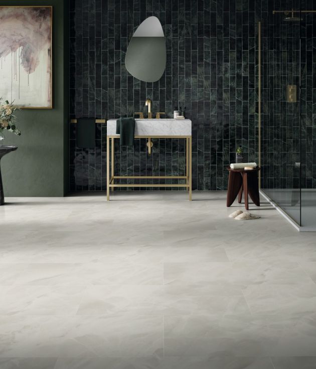 Bathroom with dark green wall with painting and side table and greenish-black tiles behind organic-shaped mirror, white and gold sink, and walk-in shower, with whitish-gray stone-look floors